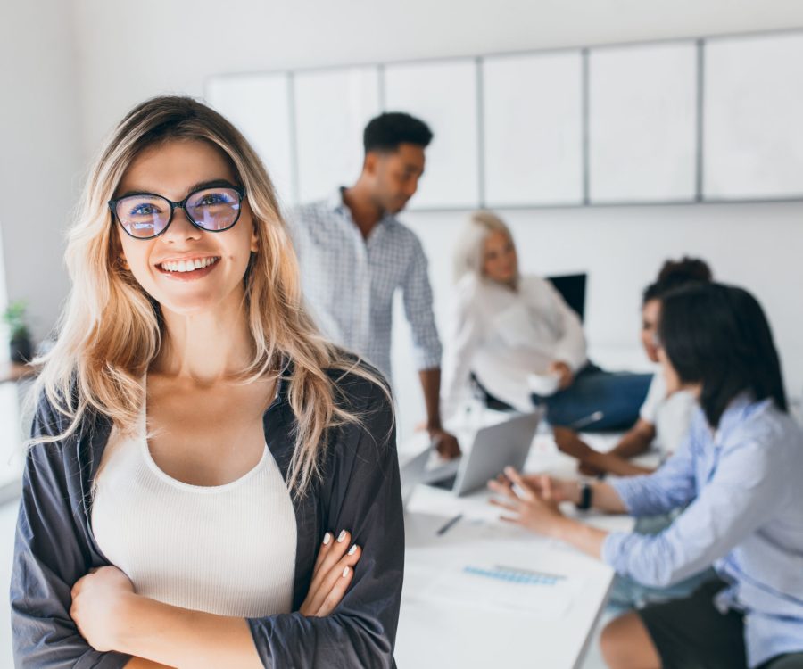 Blonde female executive posing with smile and arms crossed during brainstorm with managers. Indoor portrait of european student spending time in hall with asian and african friends.