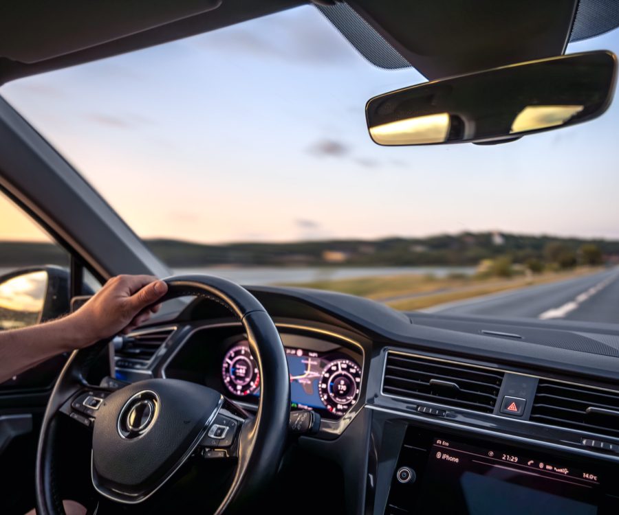 A driver's hand on the wheel during a peaceful sunset drive along a riverside highway, with dashboard navigation and glowing ambient light.